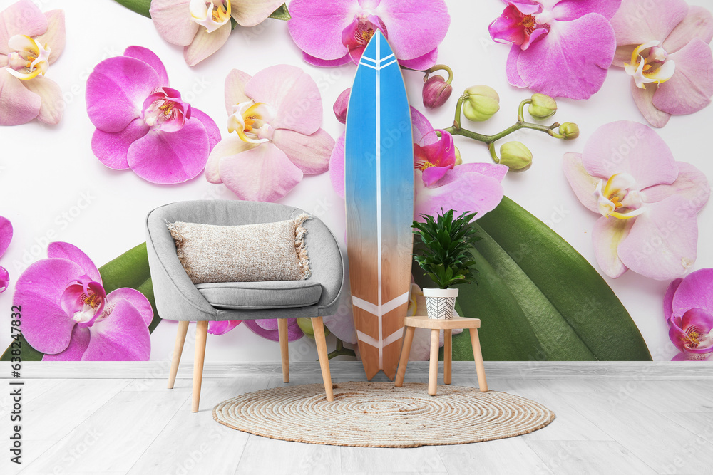 Wooden surfboard with armchair and houseplant near wall with printed orchid flowers