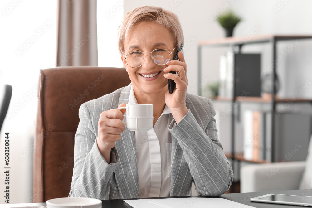 Mature businesswoman talking by phone and drinking coffee in office