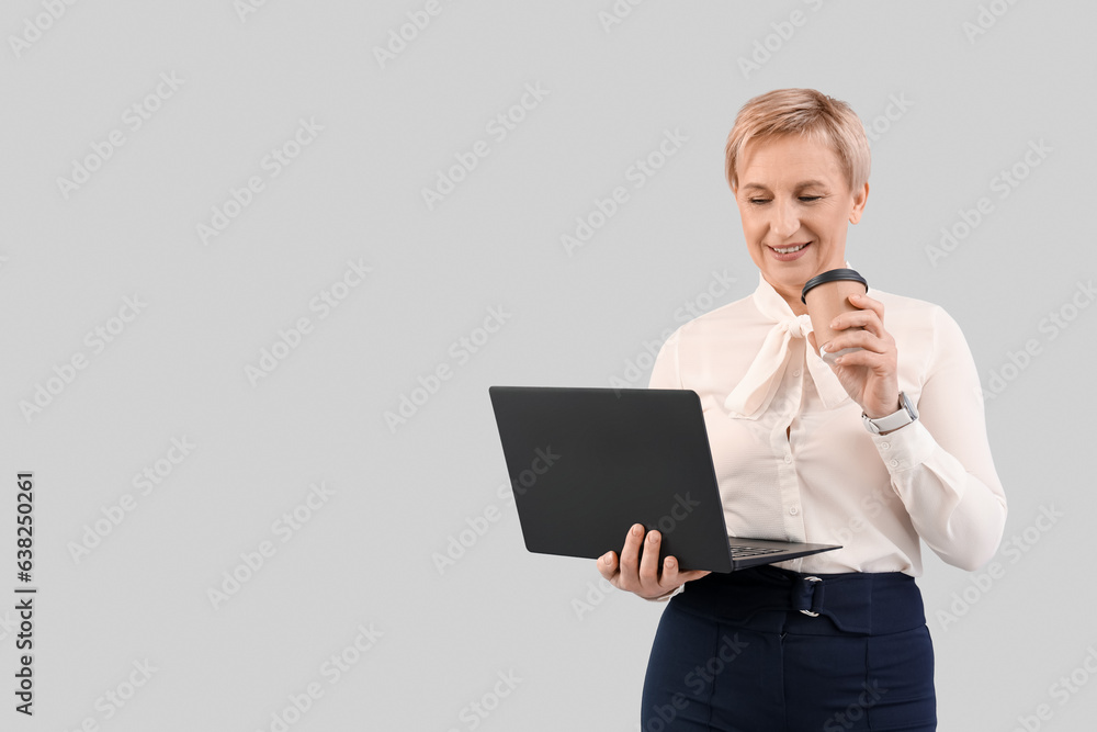 Mature businesswoman with laptop and coffee on grey background