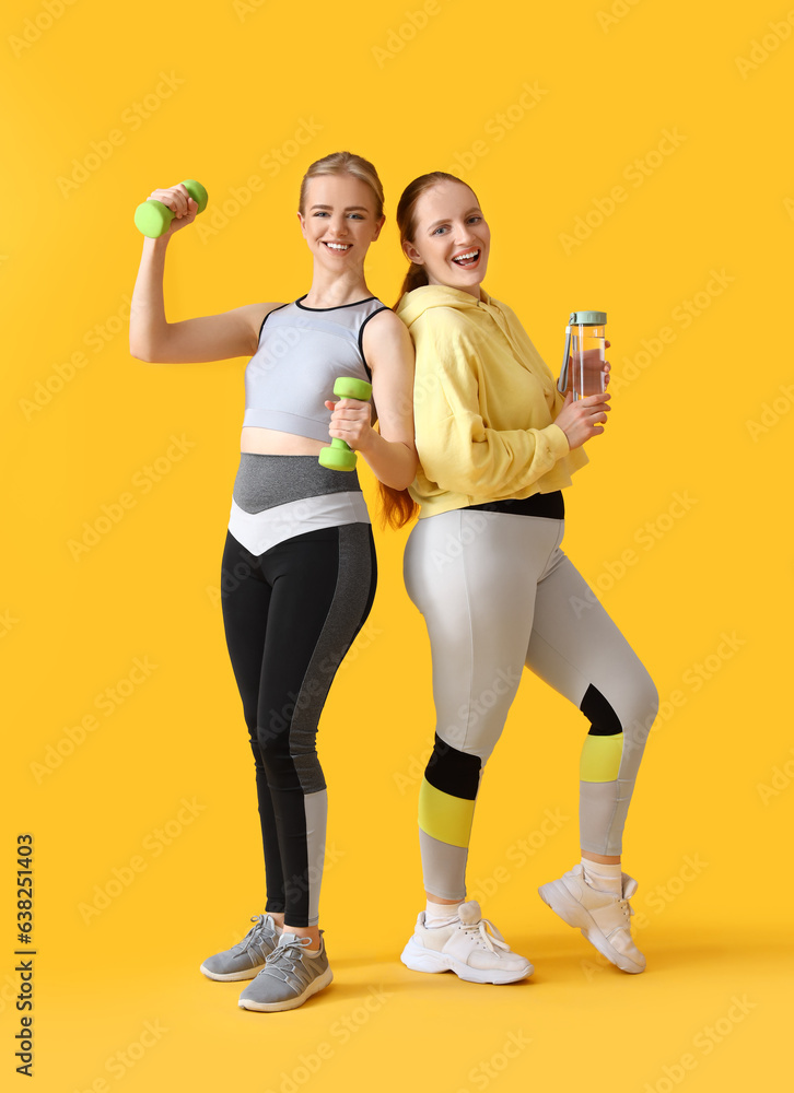 Portrait of sporty young women with dumbbells and water on yellow background