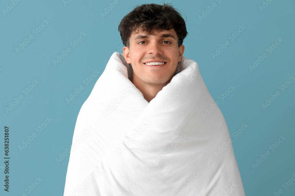 Handsome happy young man wrapped in soft blanket on blue background