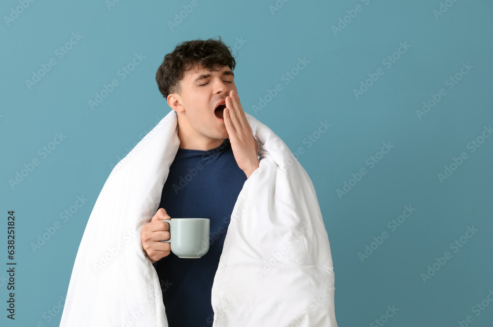 Handsome young man with cup of coffee and soft blanket yawning on blue background