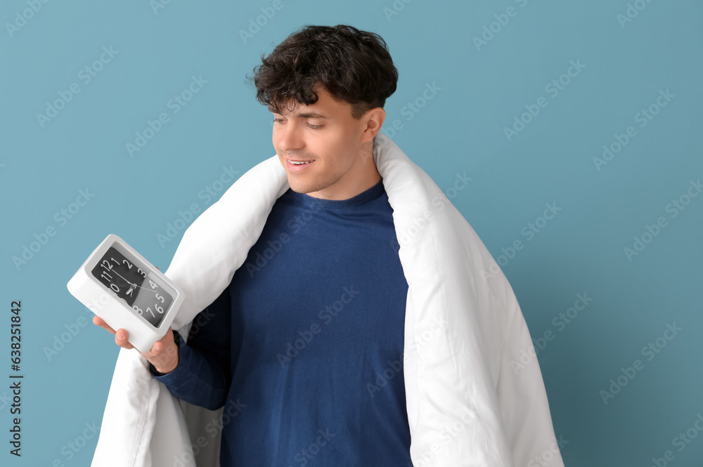 Handsome happy young man with alarm clock and soft blanket on blue background