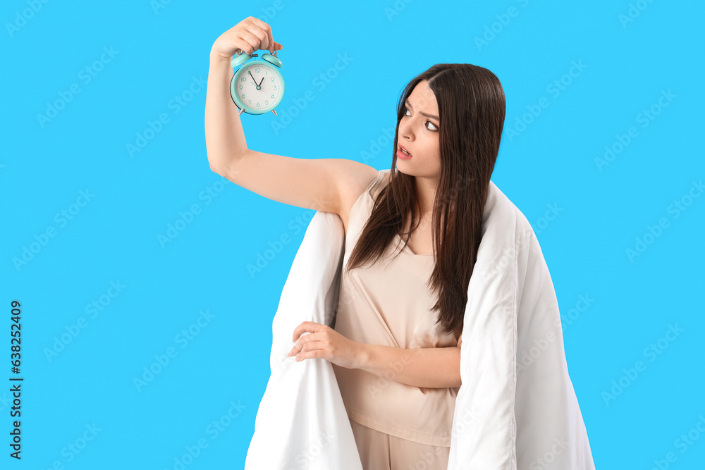 Upset young woman with alarm clock and soft blanket on blue background