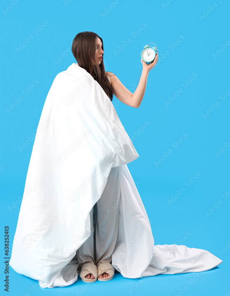 Upset young woman with alarm clock and soft blanket on blue background