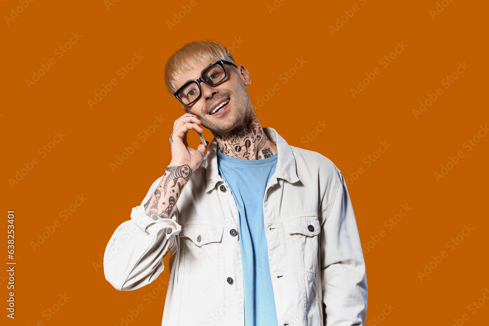 Young tattooed man in stylish eyeglasses talking by mobile phone on color background