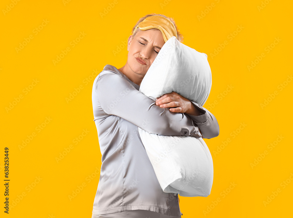 Sleepy mature woman in pajamas with pillow on yellow background