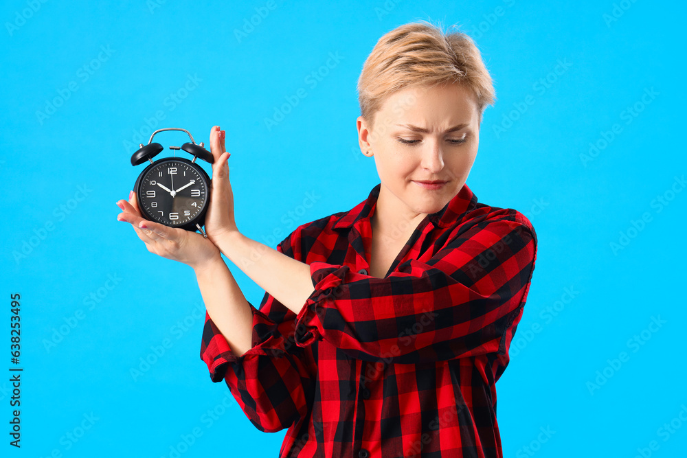 Displeased young woman in pajamas with alarm clock on blue background