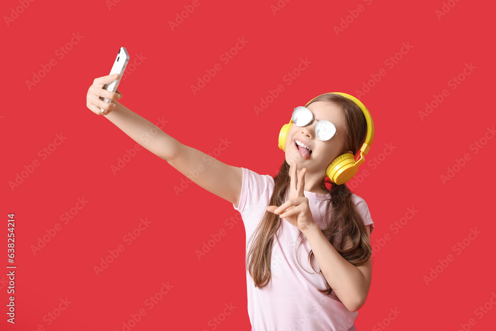 Little girl in headphones with mobile phone taking selfie on red background