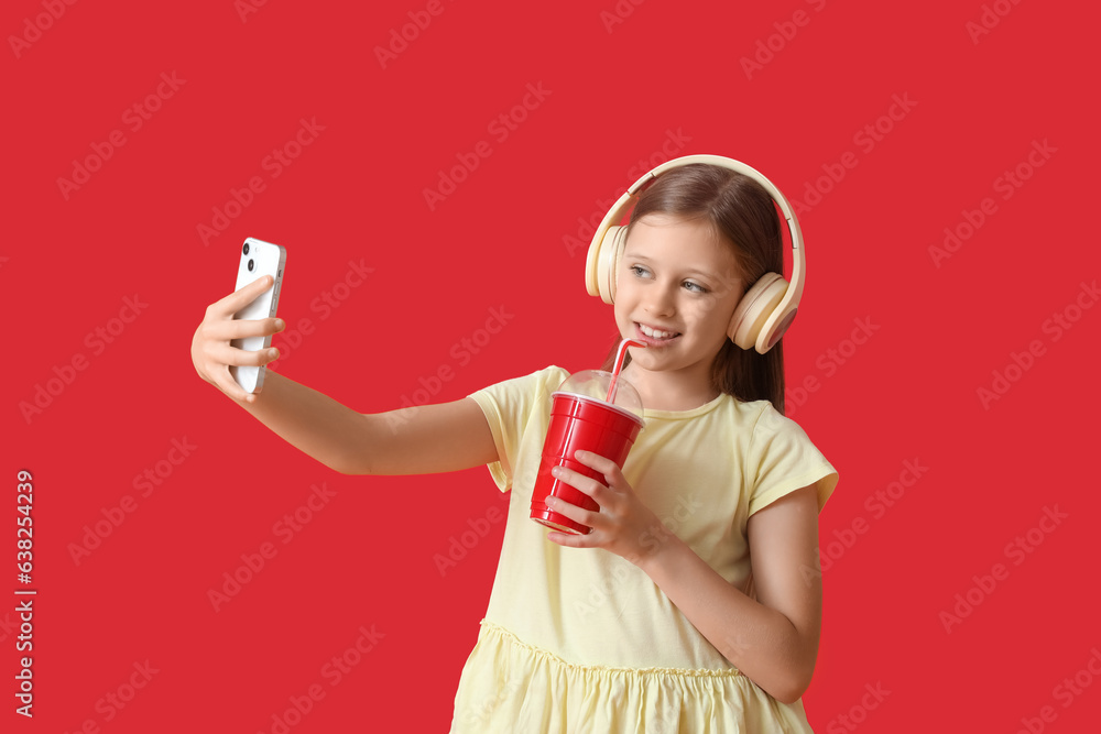 Little girl in headphones with cup of soda taking selfie on red background