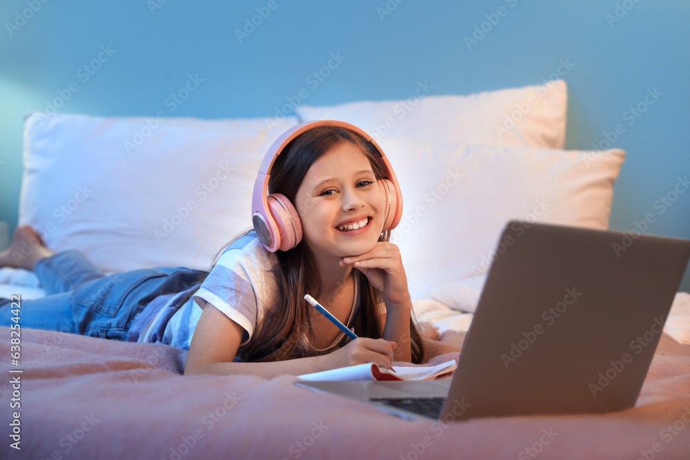 Little girl with headphones studying online in bedroom at night