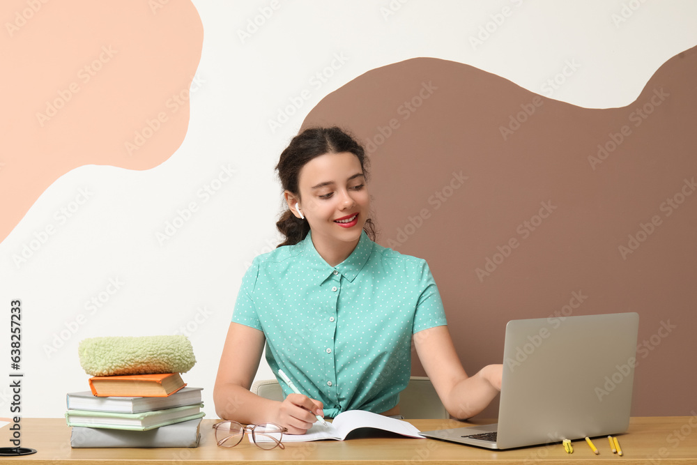 Female student in earphones with laptop doing homework at table near color wall