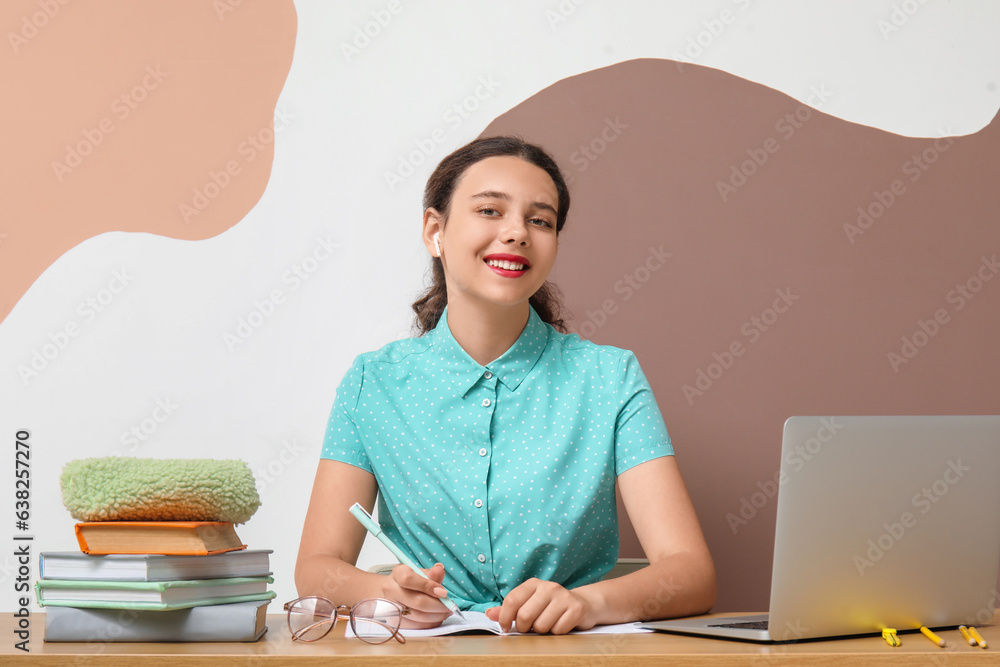 Female student in earphones with laptop doing homework at table near color wall