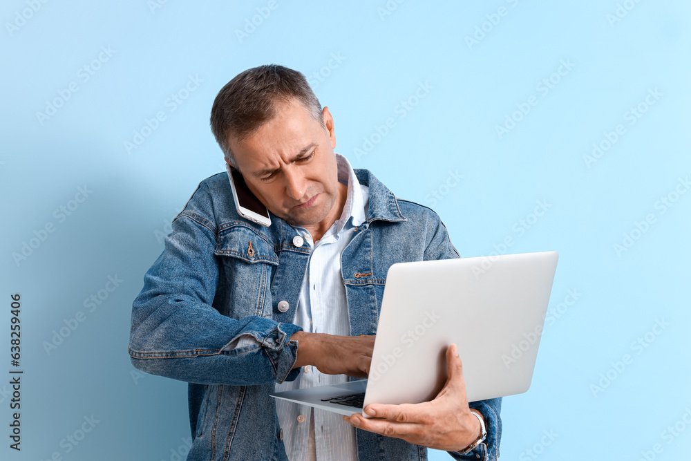 Stressed mature man with laptop talking by mobile phone on blue background. Deadline concept