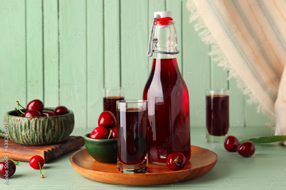 Bottle and glasses of sweet cherry liqueur with berries on green wooden table near wall