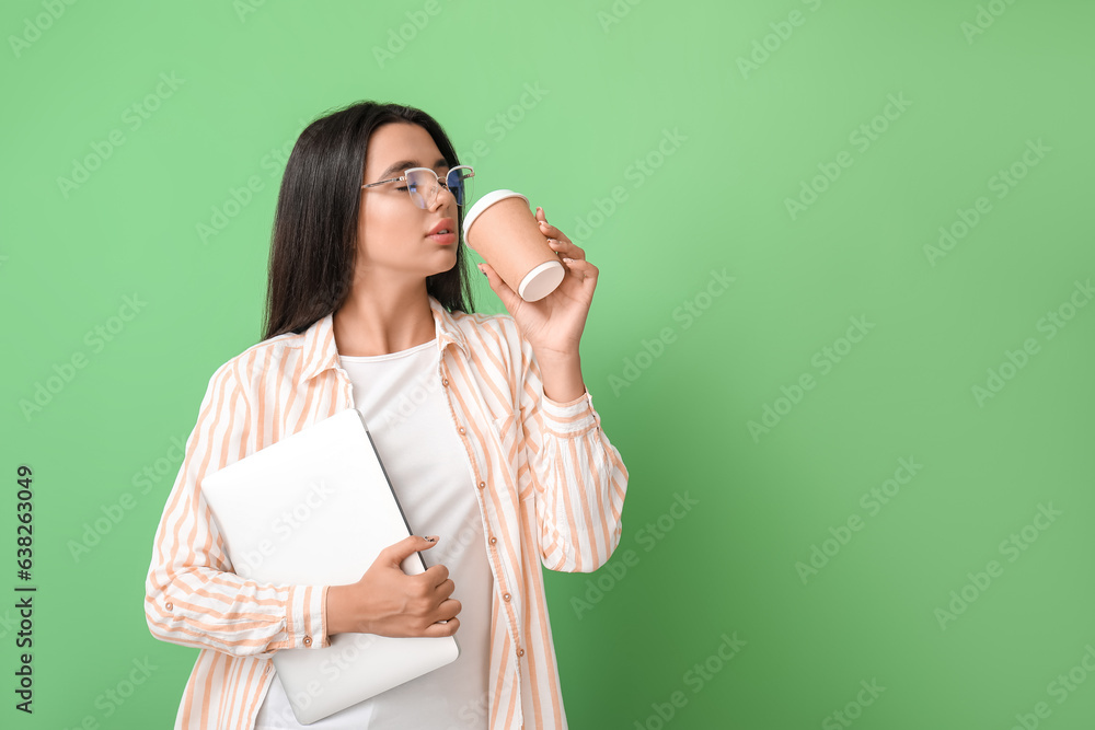 Young female programmer with laptop drinking coffee on green background