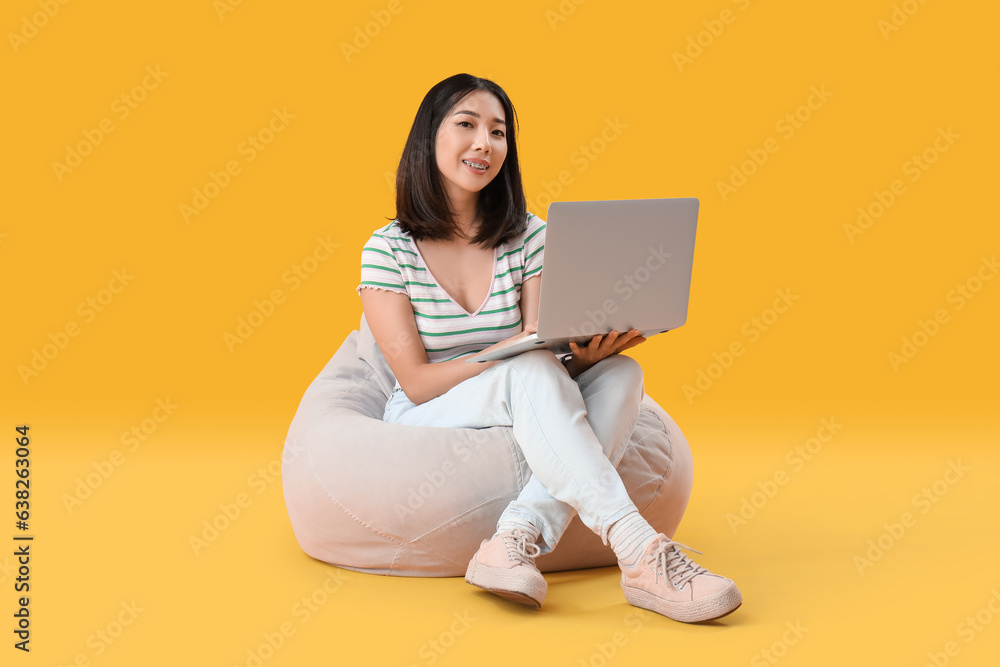 Happy young female Asian programmer with laptop sitting on beanbag chair against yellow background