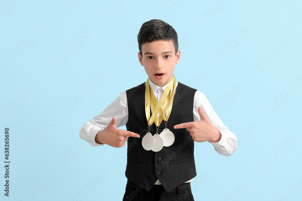 Surprised little school boy with golden medals on light blue background