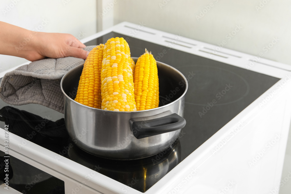 Woman boiling corn cobs in cooking pot on electric stove in kitchen