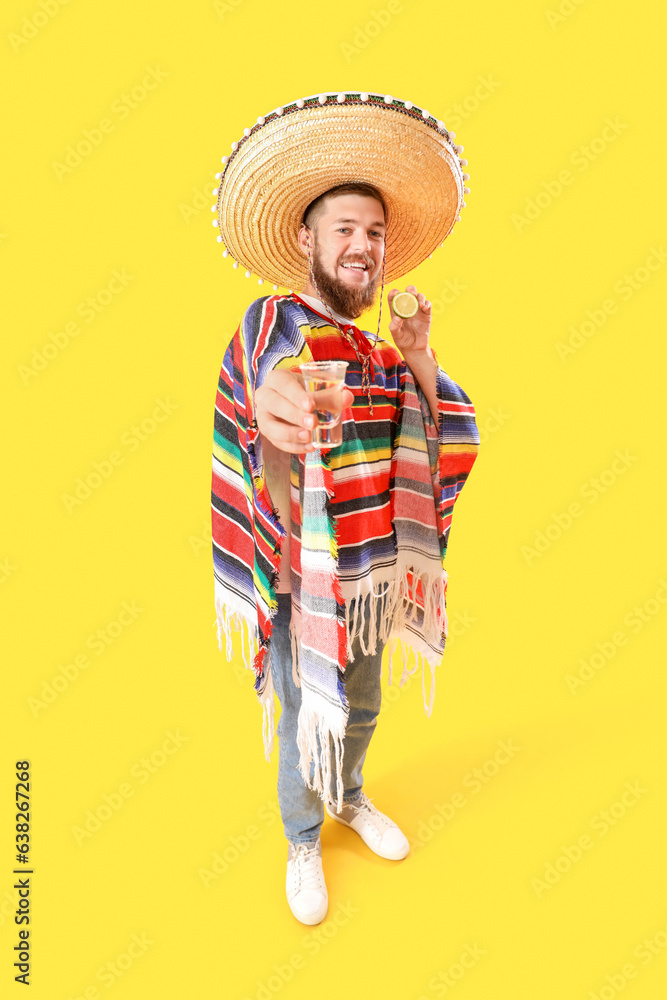 Handsome young man in Mexican poncho and sombrero holding shot of tequila on yellow background