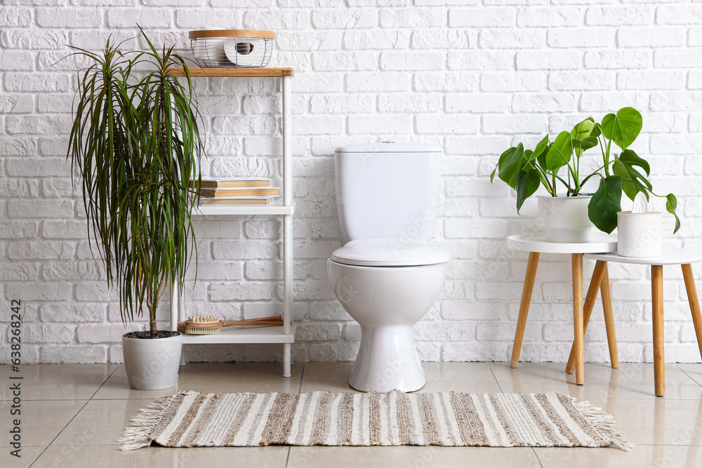 Interior of light restroom with ceramic toilet bowl, shelving unit and houseplants near white brick 