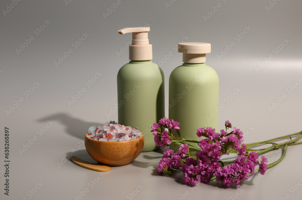 Bottles of cosmetic products with beautiful flowers and sea salt on grey background