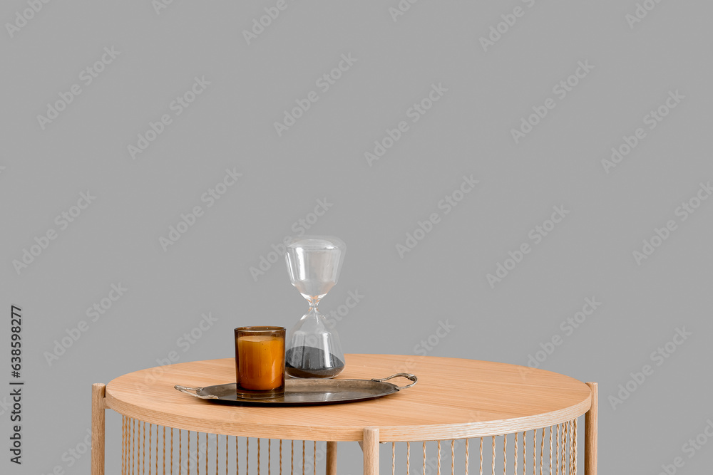 Tray with hourglass and candle on wooden coffee table near grey wall