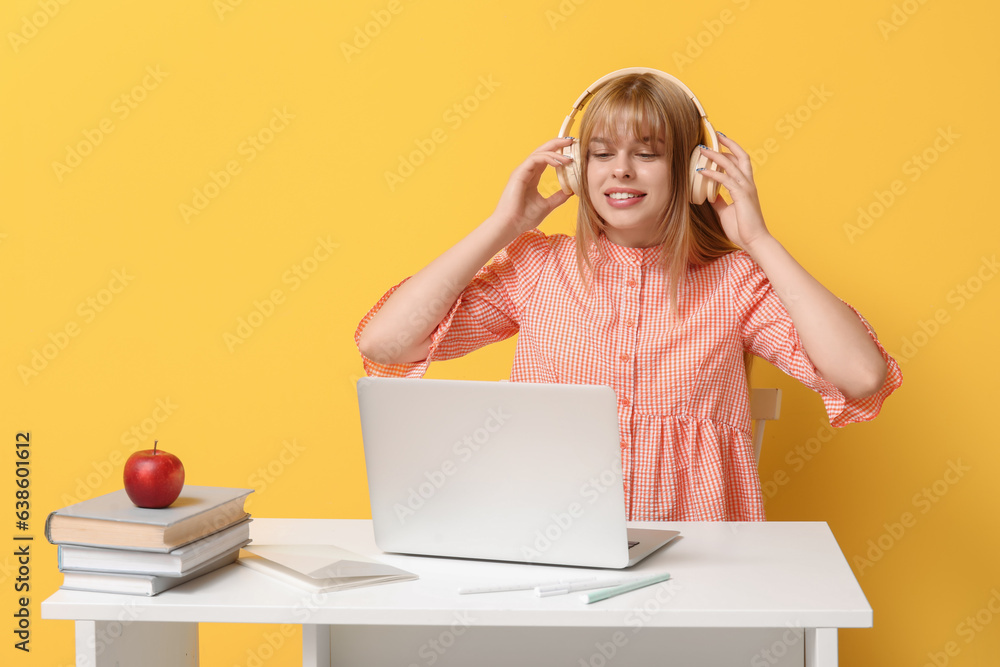 Female student in headphones doing homework at table on yellow background