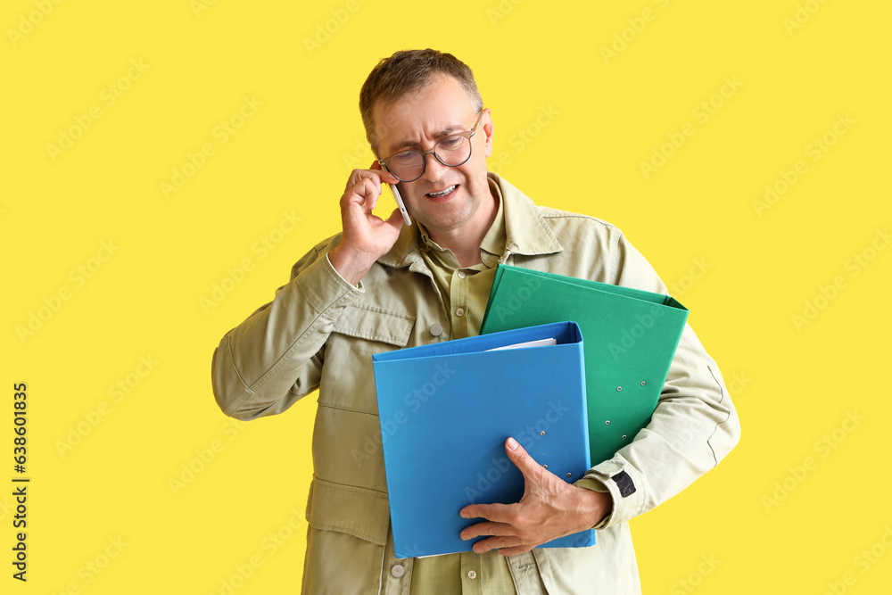 Stressed mature man with folders talking by mobile phone on yellow background. Deadline concept