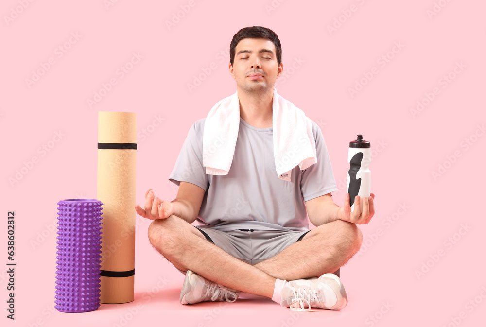 Sporty young man with bottle of water meditating on pink background