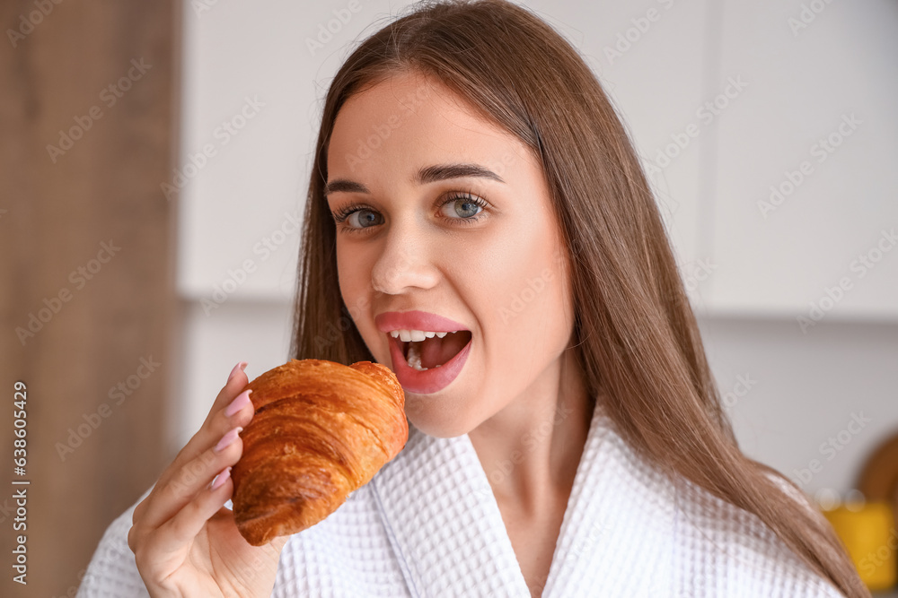 Beautiful young woman in bathrobe eating tasty croissant at kitchen