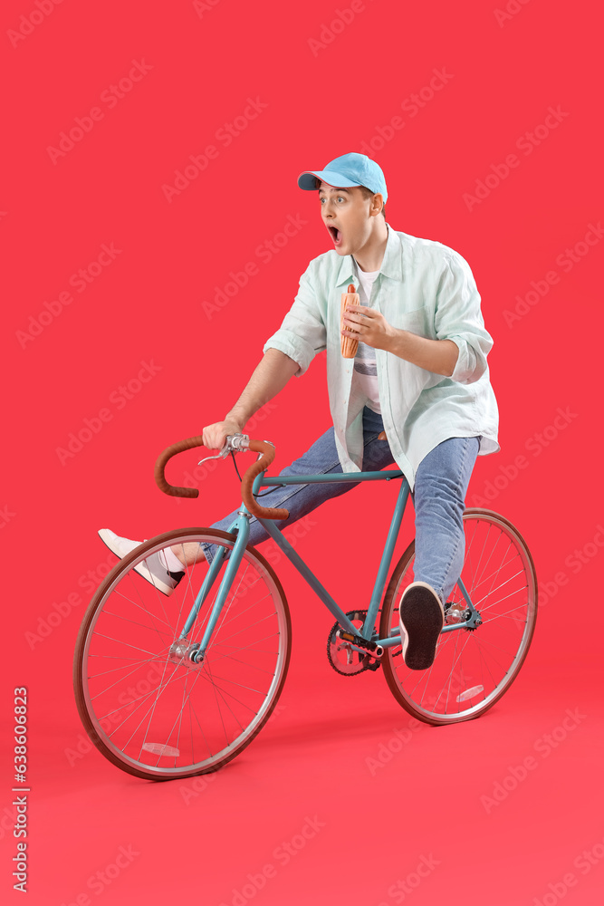 Surprised young man with tasty hot dog and bicycle on red background