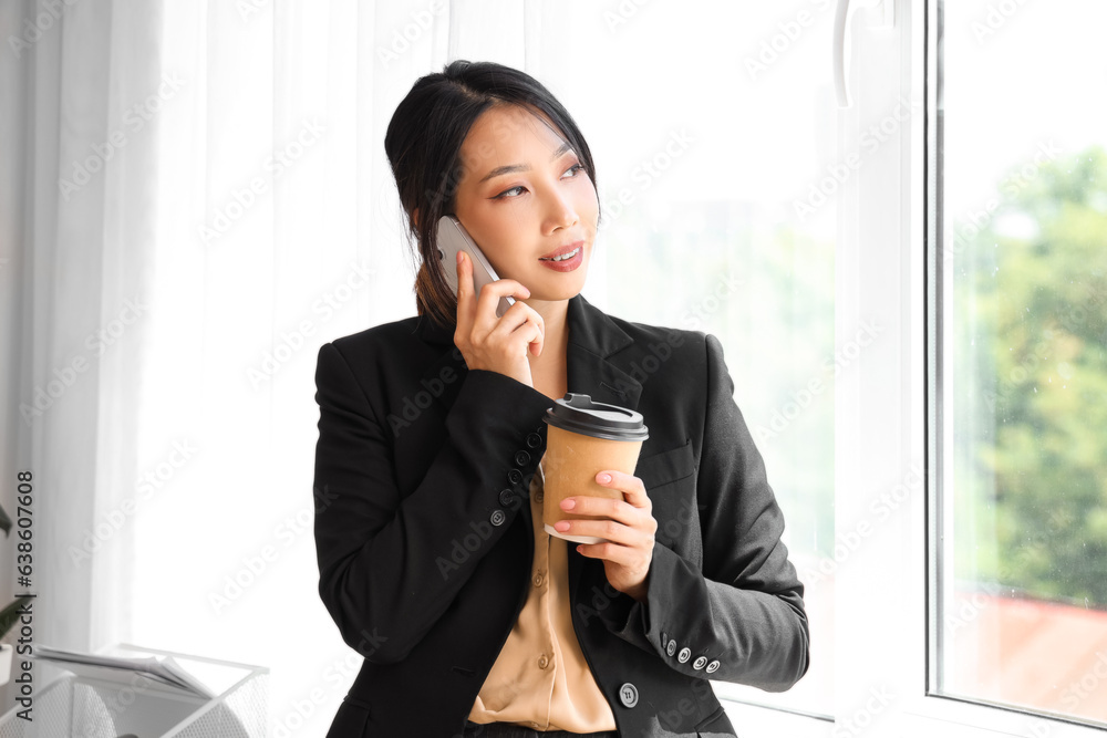 Young Asian businesswoman with cup of coffee talking by mobile phone in office