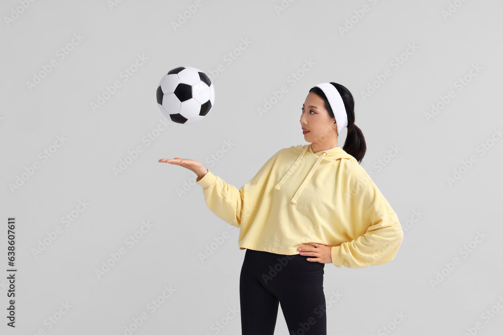 Happy young sporty Asian woman playing with soccer ball on grey background