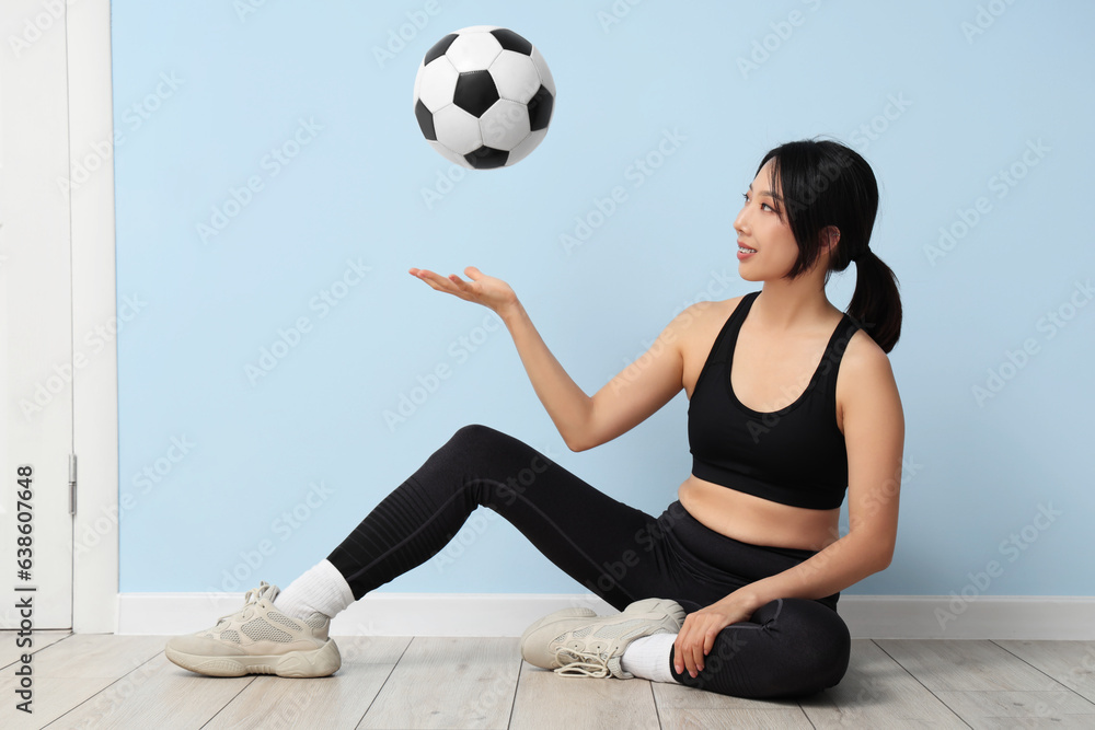 Happy young sporty Asian woman with soccer ball sitting on floor near blue wall