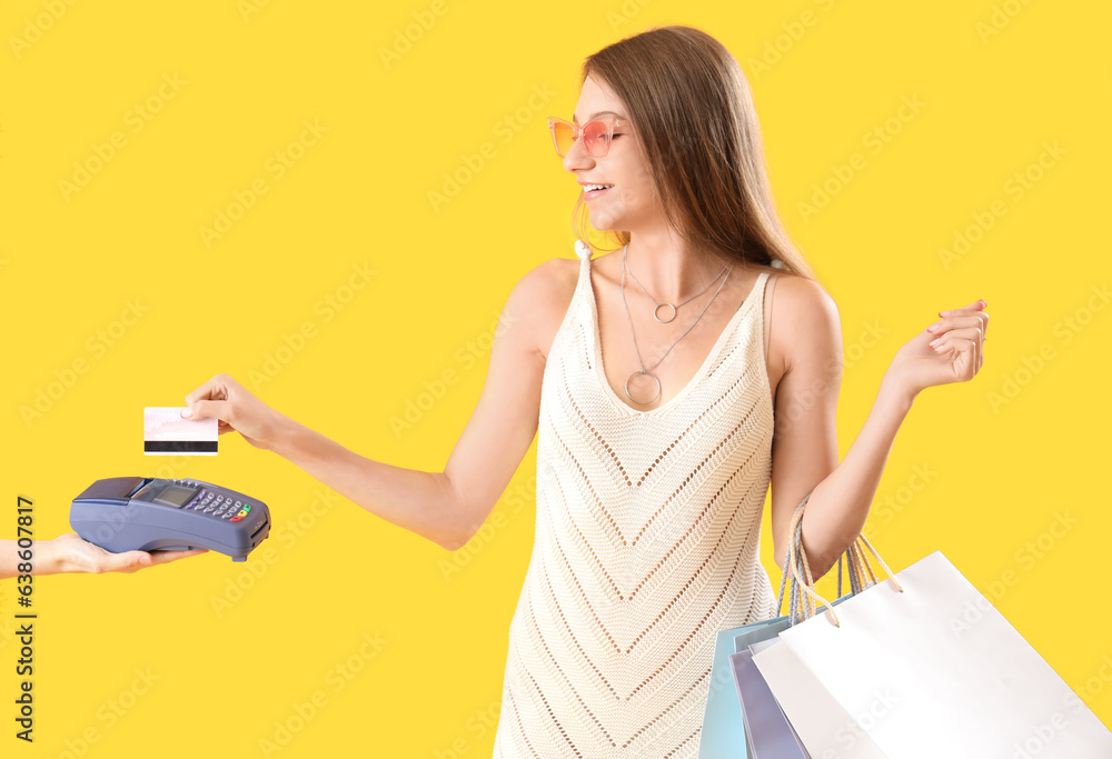 Happy stylish young woman with shopping bags and credit card paying via terminal on yellow backgroun