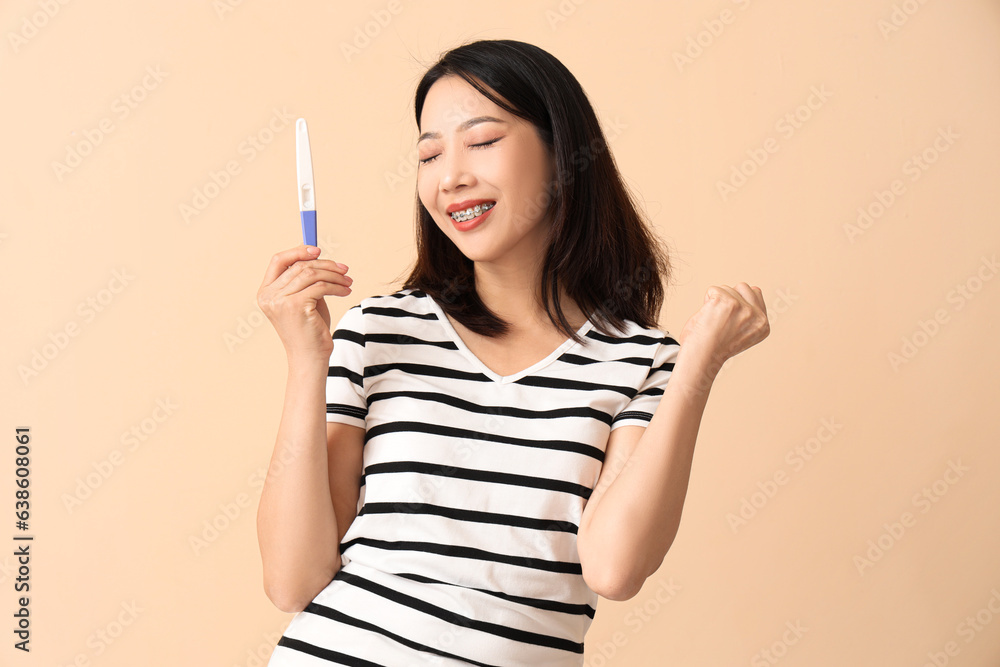 Happy beautiful young Asian woman with pregnancy test on beige background