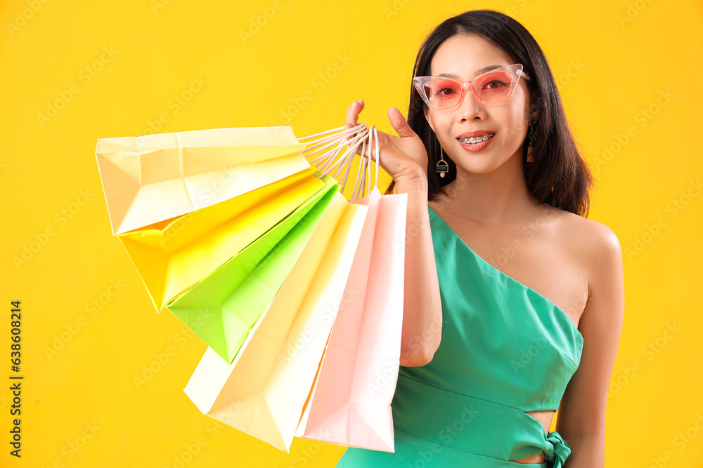 Beautiful happy stylish young Asian woman with shopping bags on yellow background