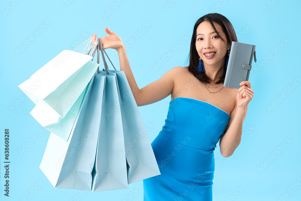 Beautiful stylish young Asian woman with shopping bags and wallet on blue background