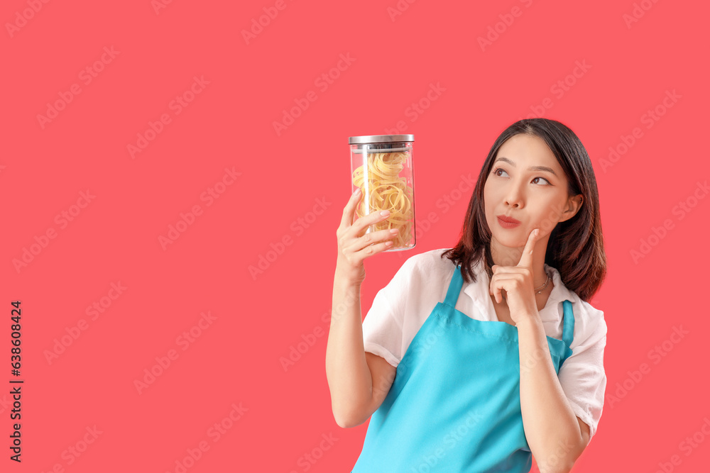 Thoughtful young beautiful Asian woman with jar of raw pasta on red background