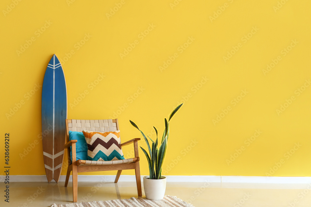 Stylish armchair, surfing board and houseplant near yellow wall