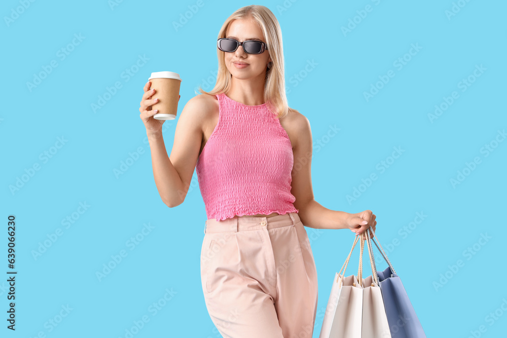 Beautiful happy young woman with cup of coffee and shopping bags on blue background