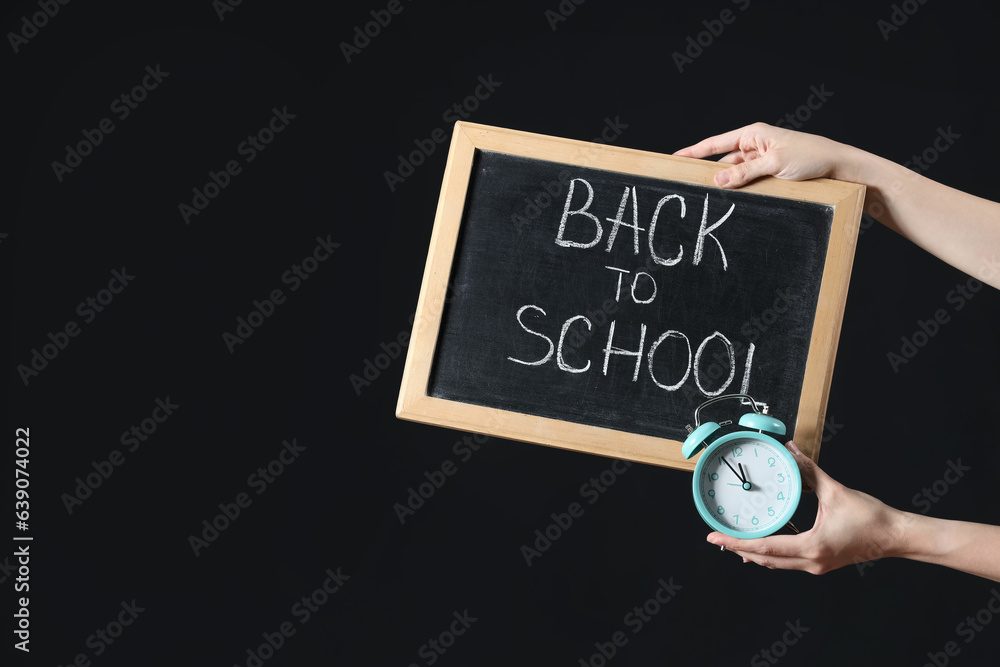 Female hands holding alarm clock and blackboard with text BACK TO SCHOOL on black background