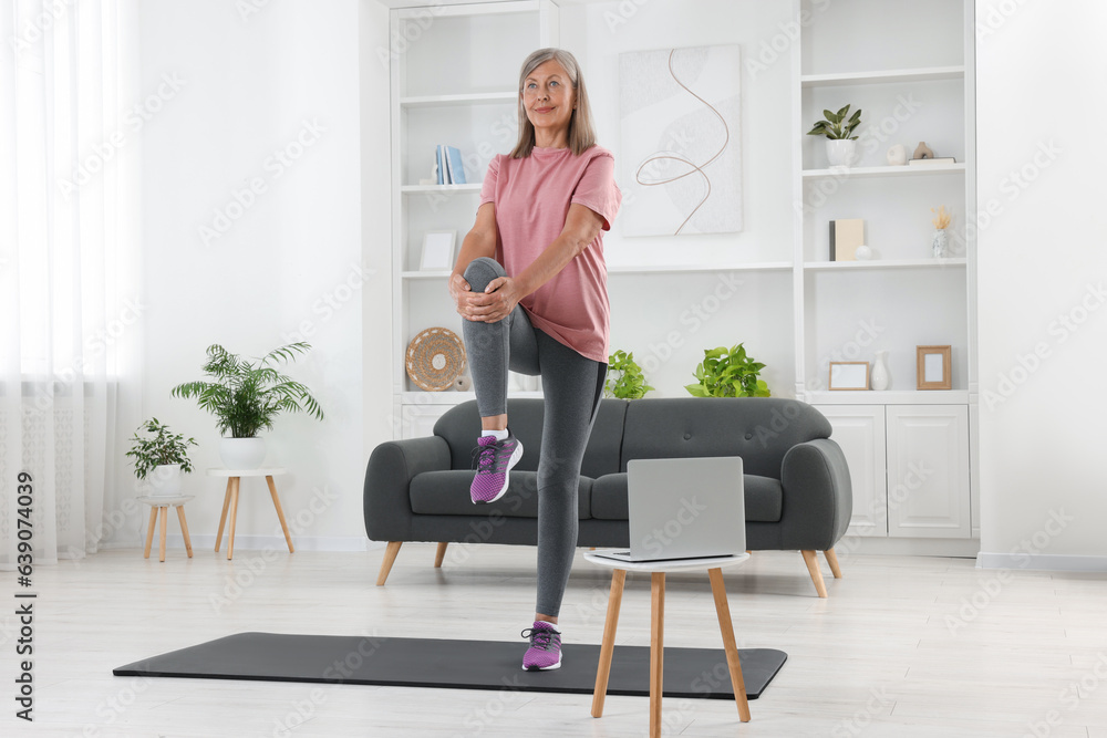 Senior woman in sportswear stretching near laptop at home