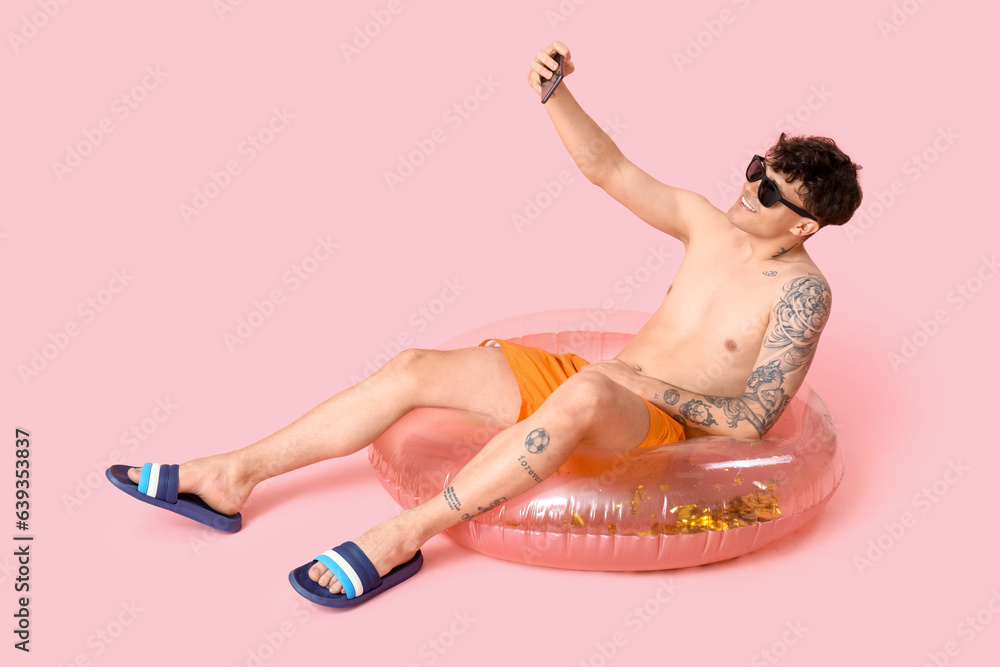 Young man with inflatable ring taking selfie on pink background