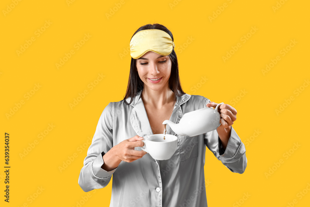 Young woman in pajamas pouring tea into cup on yellow background