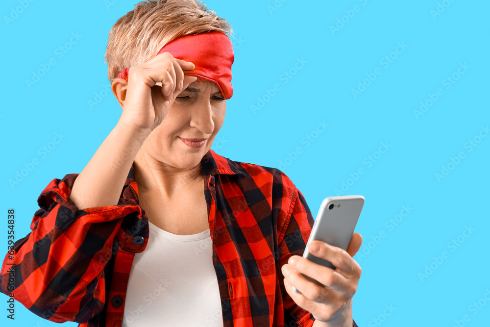 Mature woman in pajamas and sleeping mask using mobile phone on blue background