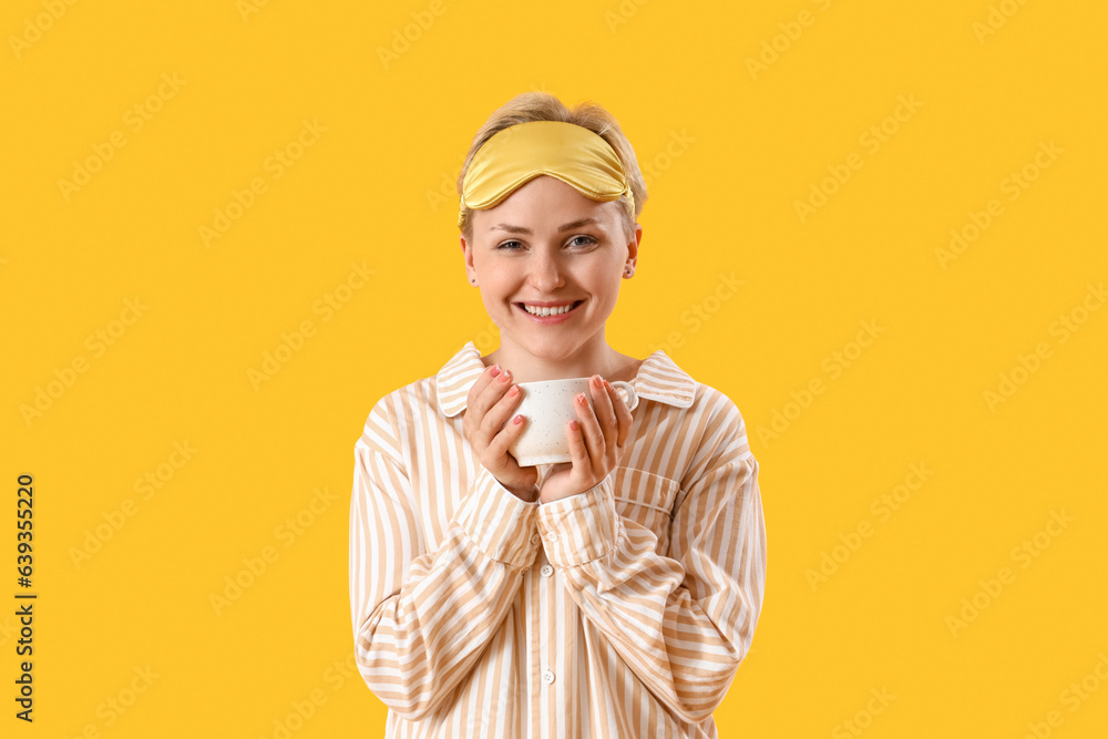 Young woman in pajamas with cup of coffee on yellow background