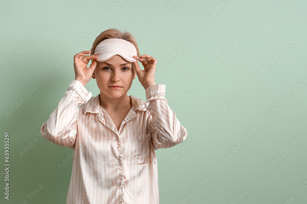 Young woman in pajamas and sleeping mask on green background