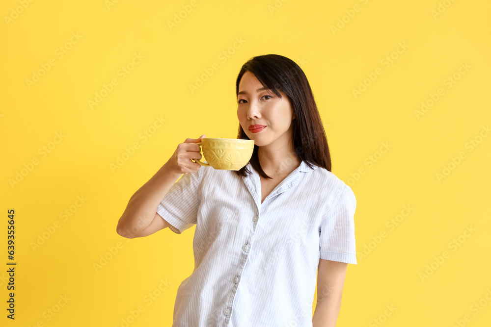 Beautiful Asian woman in pajamas with cup of coffee on yellow background